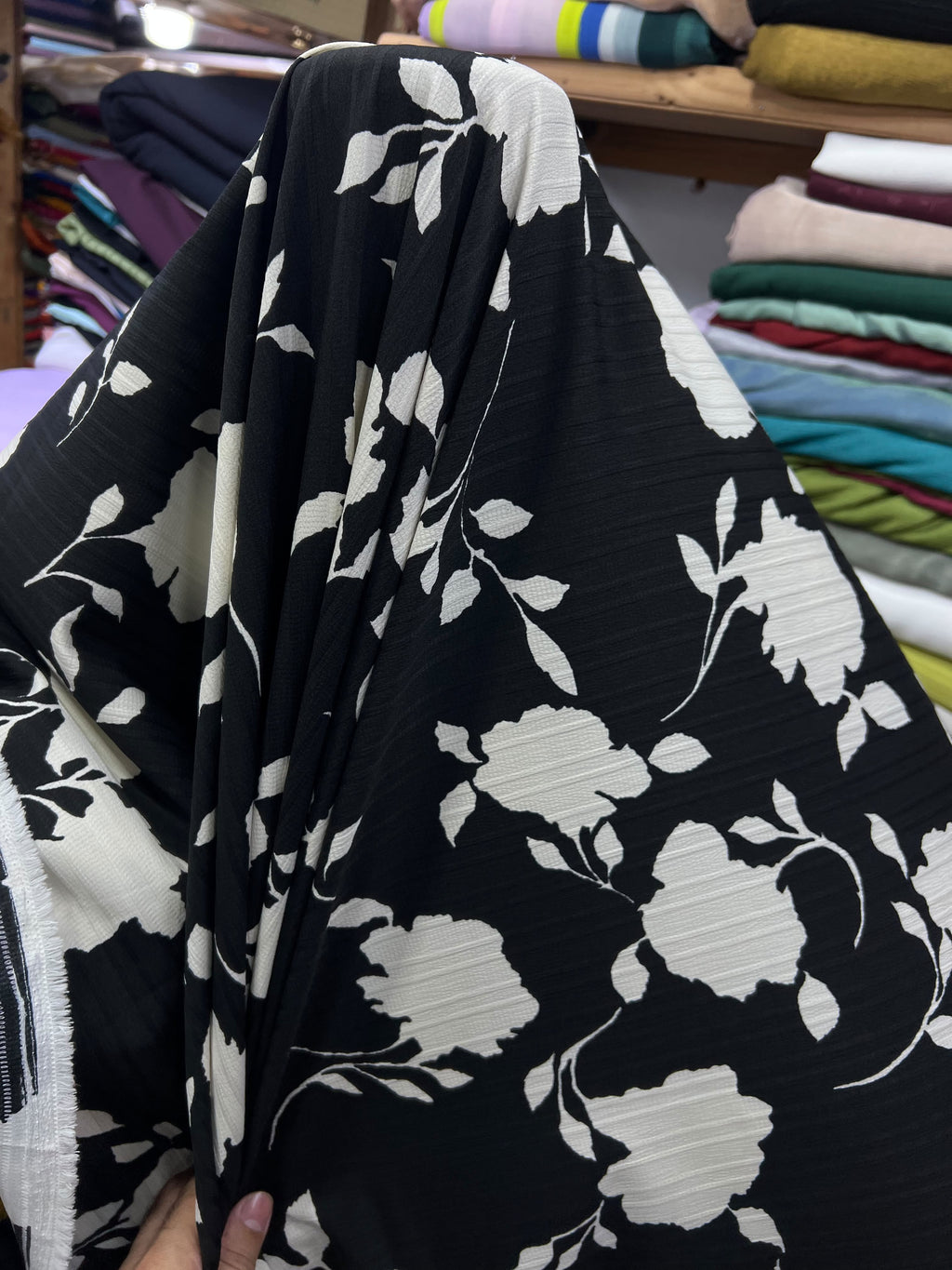A close-up of a black fabric with a large white floral and leaf pattern, held up by a hand in a fabric store with folded textiles in the background.