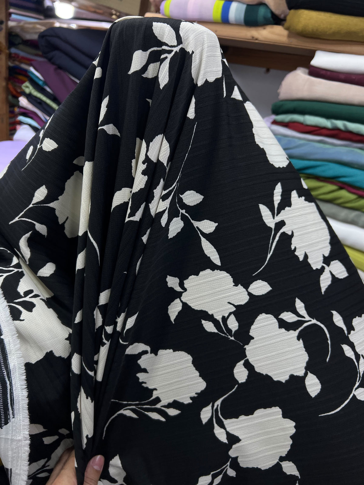 A close-up of a black fabric with a large white floral and leaf pattern, held up by a hand in a fabric store with folded textiles in the background.